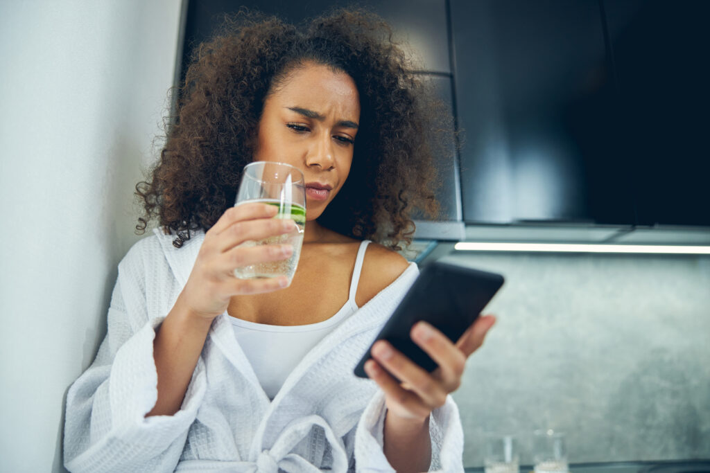 Ideges fiatal nő nézi a mobiltelefonját, kezében pohár víz