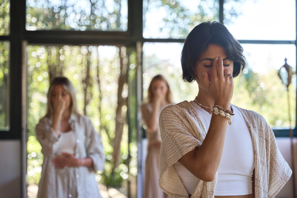 Női csoport csukott szemmel meditál, jógázik, pranayama légzőgyakorlatokat végez, ujjaikat az arcukon tartva, energiát érezve holisztikus gyógyító jógaedzés és elvonulás közben.