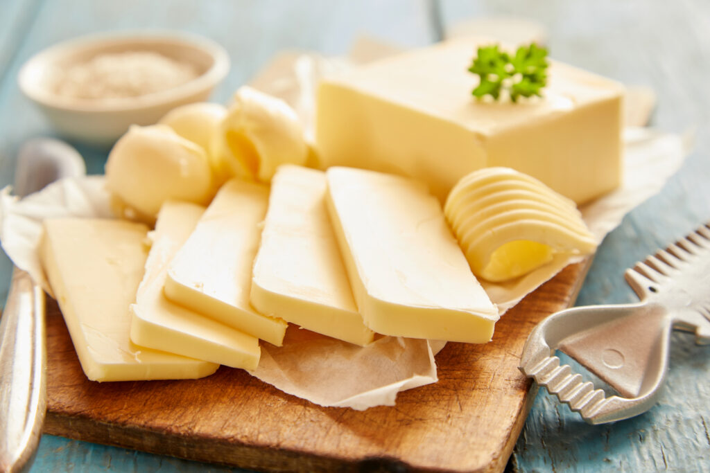 Közelről készült kép egy vágódeszkán szeletelt vajról, a háttérben kék fa.