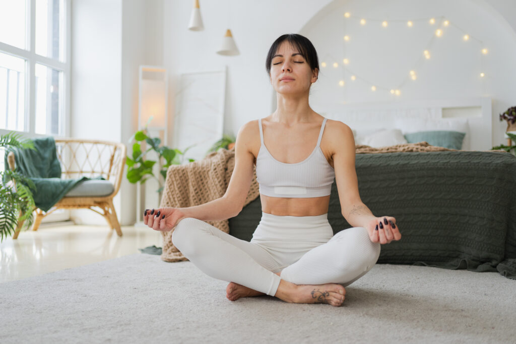 Jóga mindfulness meditáció. Fiatal, egészséges nő jógázik otthon a nappaliban. Nő lótuszpózban ül, meditál, mosolyog, pihentető beltéri környezetben. Lány légzőgyakorlatokat végez. Jóga otthon