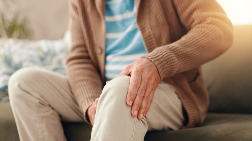Idősebb férfi, kanapé és térdfájdalom krónikus betegség, ízületi gyulladás vagy fibromyalgia miatti sérüléssel otthon. Közeli kép: idős vagy férfi személy fájó lábbal, baleset vagy ízületi gyulladás miatti orvosi vészhelyzet.