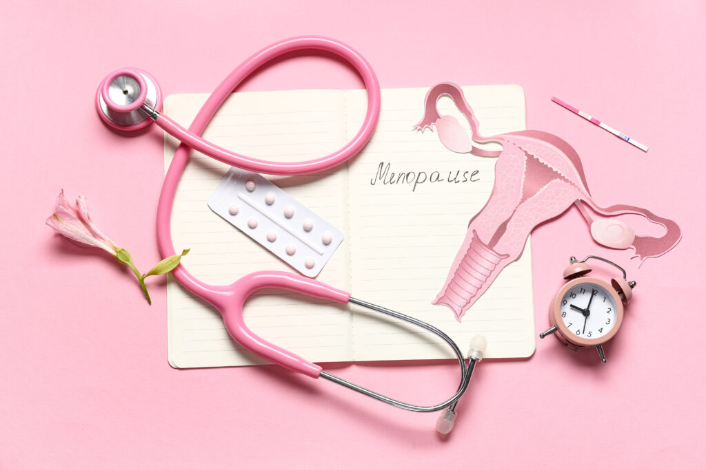 Jegyzetfüzet a MENOPAUZA szóval, tabletták, sztetoszkóp és papírméhtest rózsaszín háttéren