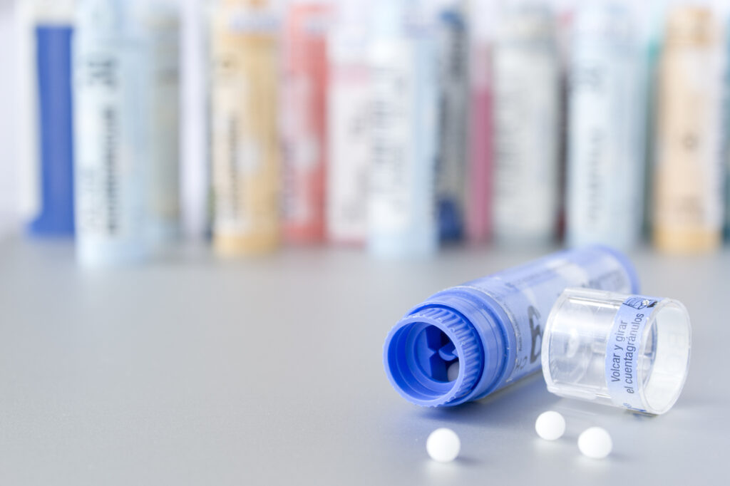 Egy kék homeopátiás gyógyszeres doboz nyitva egy alumínium felületen. Három fehér tabletta rajta. Kék doboz. A háttérben további elmosódott homeopátiás gyógyszeres dobozok láthatók.