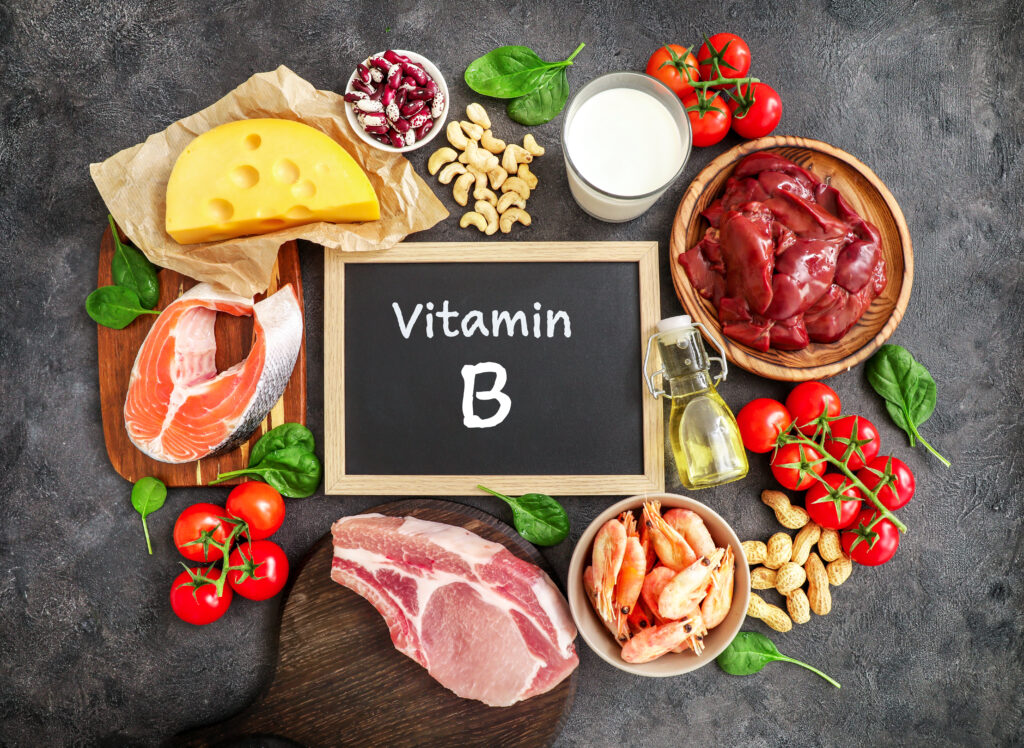 Magas B-vitamin tartalmú termékek választéka sötét háttér előtt: tej, máj, olívaolaj, paradicsom, garnélarák, földimogyoró, marhahús, spenót, lazac, kesudió, sajt, fehérbab. Felülnézet.