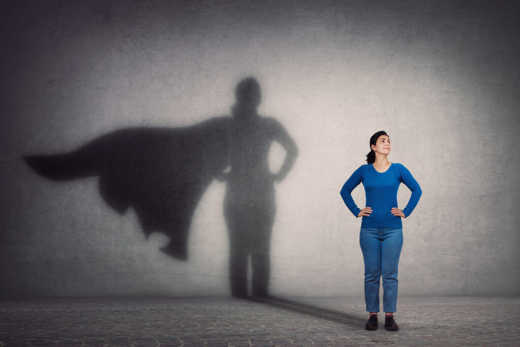 Fiatal kék ruhás nő áll egy szürke fal előtt, az árnyéka pedig super woman-ként mutatja őt.